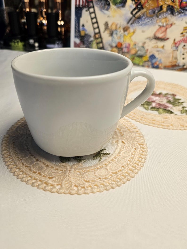 May include: A white ceramic teacup rests on a beige lace doily with a floral pattern. The doily is on a white surface. The background features a colourful illustration and blurred lights.