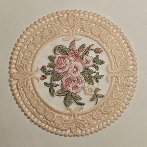 Può includere: Una tovaglietta rotonda ricamata con un bouquet floreale al centro. I fiori sono rosa e bianchi con foglie verdi. La tovaglietta ha un delicato bordo in pizzo traforato di colore beige chiaro.