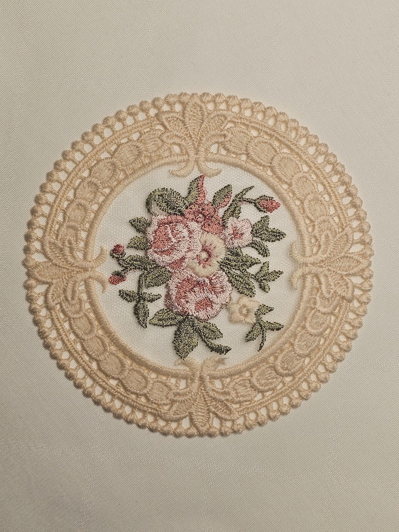 May include: A circular embroidered doily with a floral design. The central bouquet features pink and white flowers with green foliage. The doily has a delicate, openwork lace border in a light beige colour.