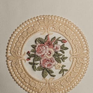 May include: A cream-coloured embroidered doily featuring a central floral bouquet of pink and white roses with green leaves. The bouquet is surrounded by a detailed, openwork lace border in a matching cream hue. The background is a plain, off-white fabric.