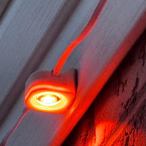 Puede incluir: Un pequeño aplique de luz cuadrado blanco con un borde rojo y una luz naranja brillante que brilla desde el centro. La luz está montada en una pared blanca.