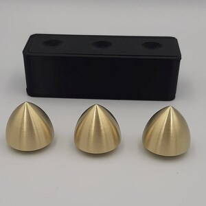 Puede incluir: Tres objetos de metal de forma c&oacute;nica de color dorado, cada uno con un acabado liso y brillante. Los objetos est&aacute;n dispuestos en fila sobre una superficie blanca. Un bloque rectangular negro se encuentra detr&aacute;s de los objetos.