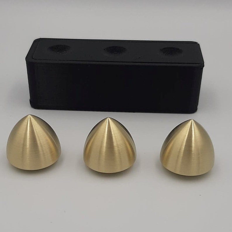 Puede incluir: Tres objetos de metal de forma c&oacute;nica de color dorado, cada uno con un acabado liso y brillante. Los objetos est&aacute;n dispuestos en fila sobre una superficie blanca. Un bloque rectangular negro se encuentra detr&aacute;s de los objetos.