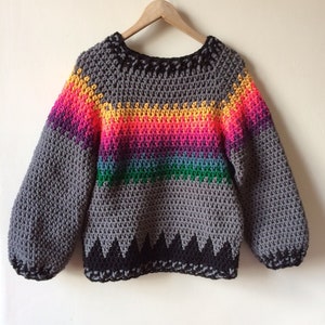 Puede incluir: Un suéter de crochet gris con una franja de arcoíris y ribete negro. El suéter tiene un corte holgado y mangas largas.