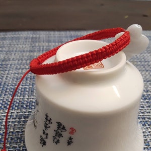 May include: A red string bracelet with a white heart charm. The bracelet is tied around a white ceramic cup.