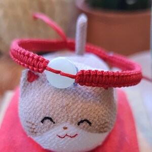May include: A small, white cat figurine with a red string bracelet around its neck. The figurine is sitting on a red cushion and a wooden box.