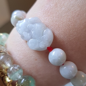 May include: A bracelet featuring smooth, pale white jade beads strung on a thin red cord, centered with a carved jade Pixiu charm. The bracelet is worn alongside several other beaded bracelets made of various translucent and multi-colored stones.