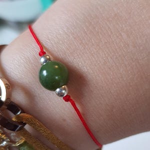 May include: A red string bracelet with a green jade bead and silver accents. Several other bracelets with gold and brown tones are visible in the background. The bracelet is a simple, elegant piece of jewelry.