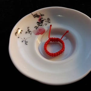 May include: A red string bracelet with a knotted design, resting on a white ceramic plate with a floral pattern.