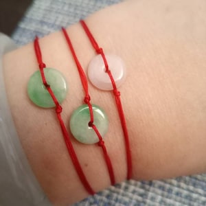 May include: Three red string bracelets with jade and pink round charms. The bracelets are tied on a person's wrist.