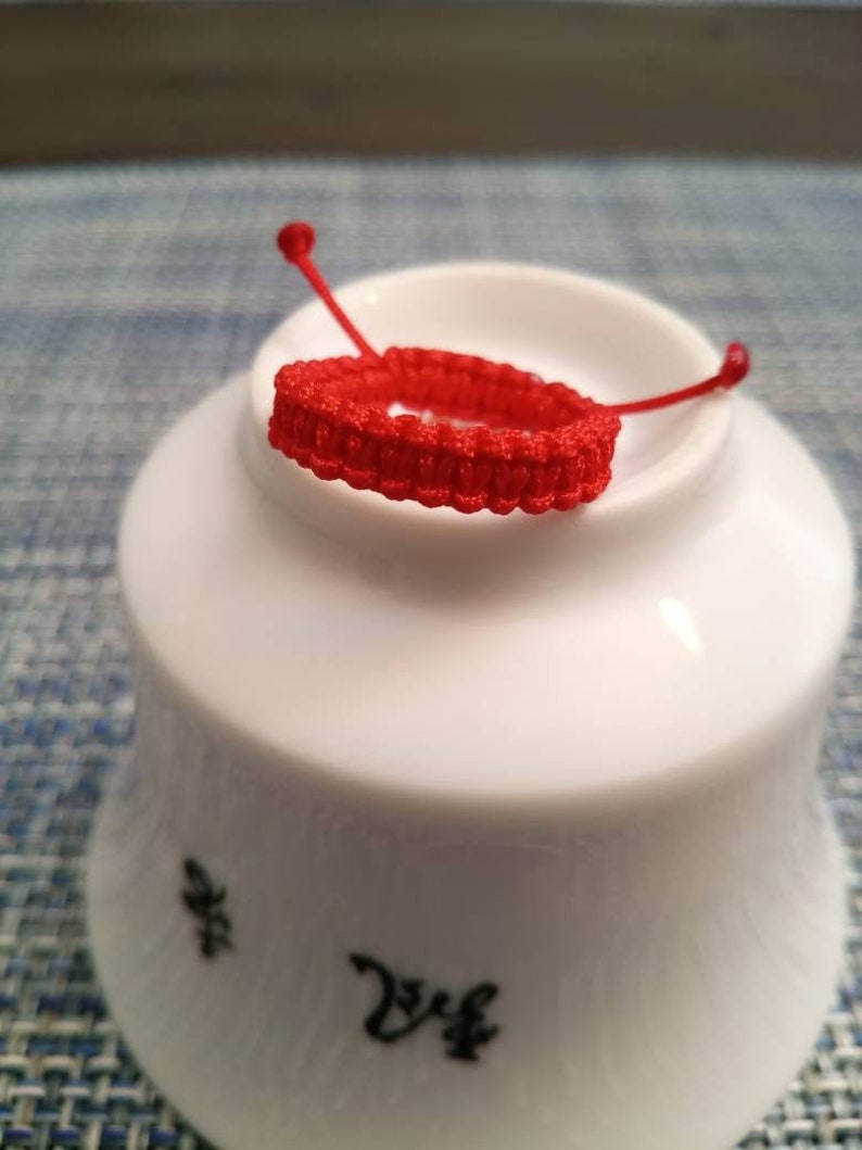 May include: A red braided string bracelet on a white ceramic dish. The dish has black writing on the bottom.