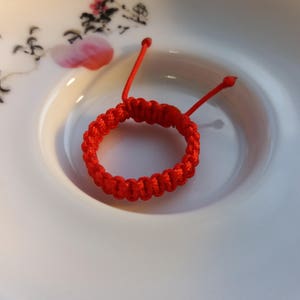 May include: A red string bracelet with a knotted design, resting on a white ceramic plate with a floral pattern.