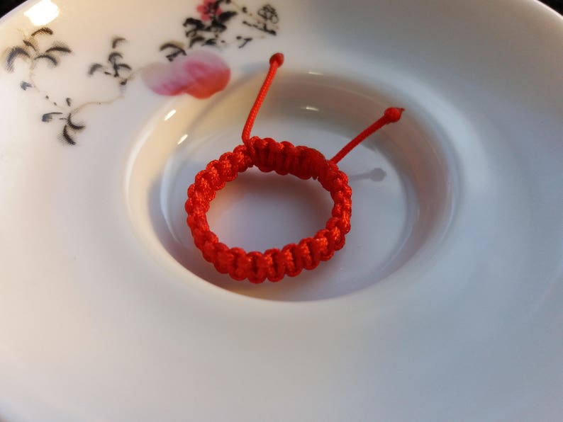 May include: A red string bracelet with a knotted design, resting on a white ceramic plate with a floral pattern.