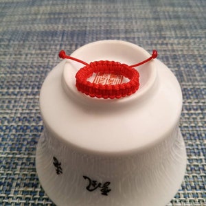 May include: A red string bracelet with a knot design. The bracelet is on a white ceramic bowl with Chinese characters.