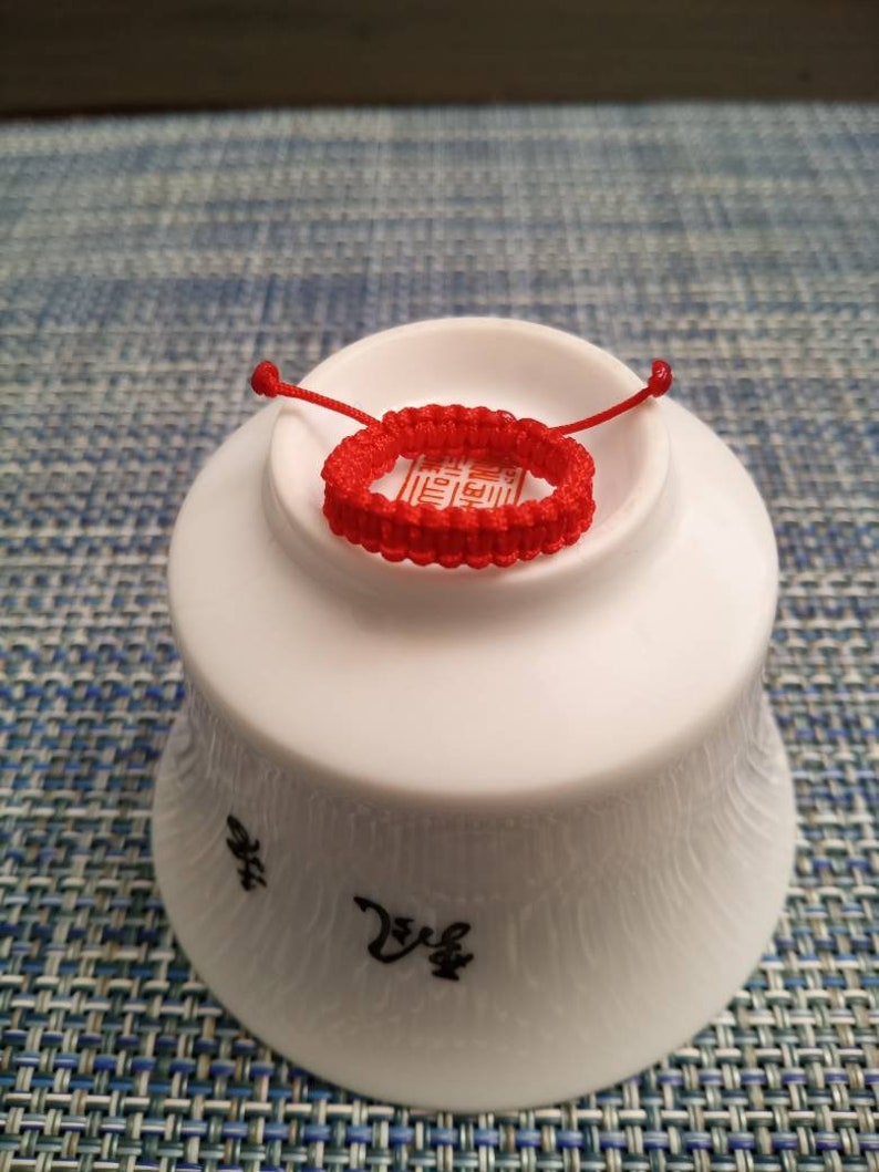 May include: A red string bracelet with a knot design. The bracelet is on a white ceramic bowl with Chinese characters.