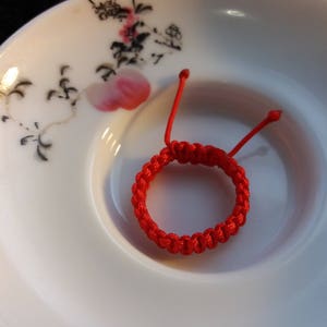 May include: A red string bracelet with a knotted design. The bracelet is on a white plate with a floral pattern.