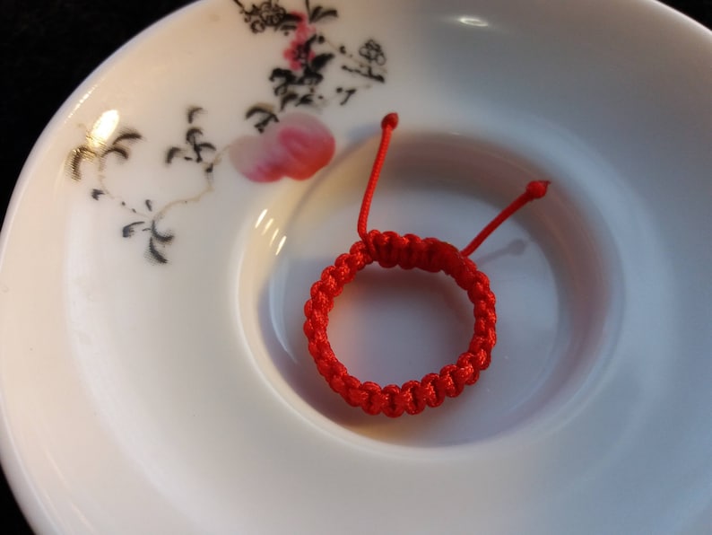 May include: A red string bracelet with a knotted design. The bracelet is on a white plate with a floral pattern.