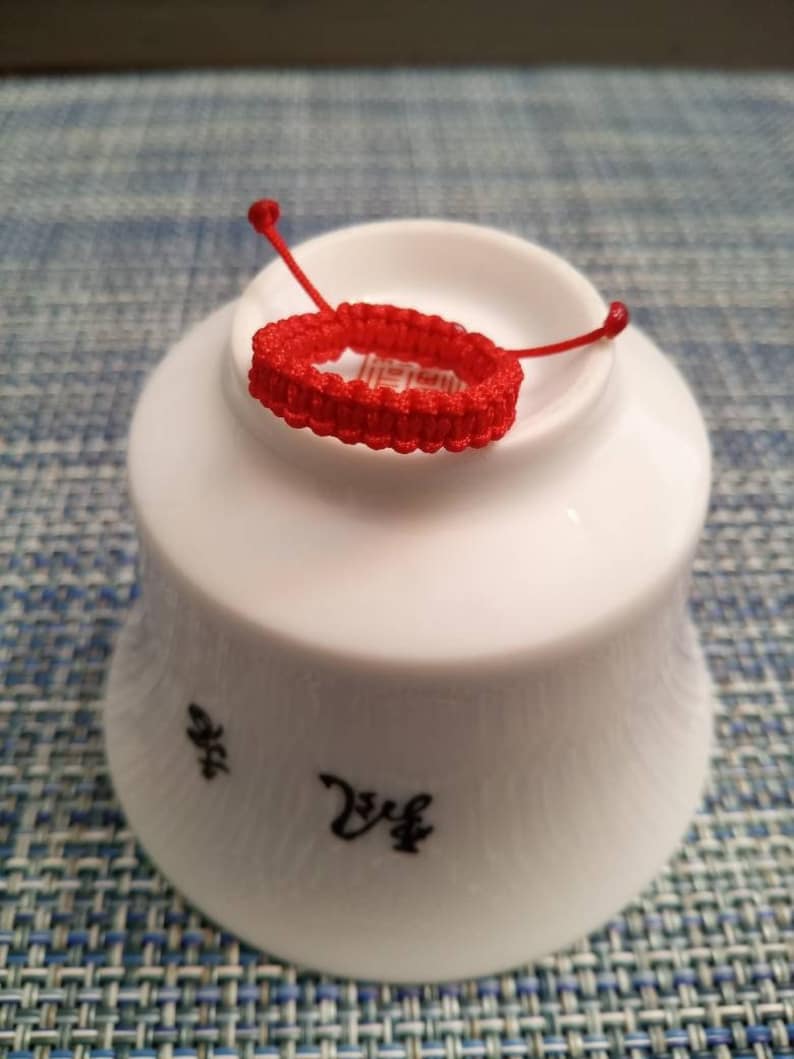 May include: A red string bracelet with a knot design, resting on a white ceramic bowl with black Chinese characters.