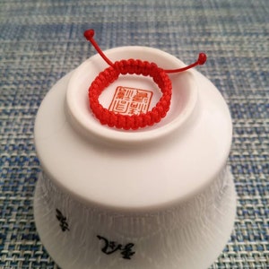 May include: A red string bracelet with a Chinese character on it. The bracelet is sitting on a white bowl.