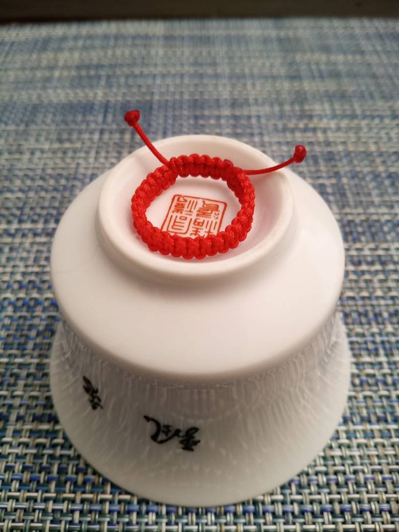 May include: A red string bracelet with a Chinese character on it. The bracelet is sitting on a white bowl.