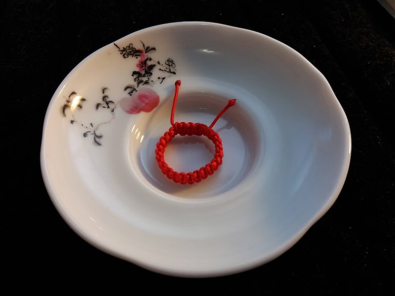 May include: A red string bracelet with a knotted design, resting on a white ceramic plate with a floral pattern.