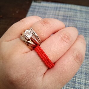 May include: A hand wearing three silver rings and a red string bracelet. The middle ring features a large, clear gemstone.