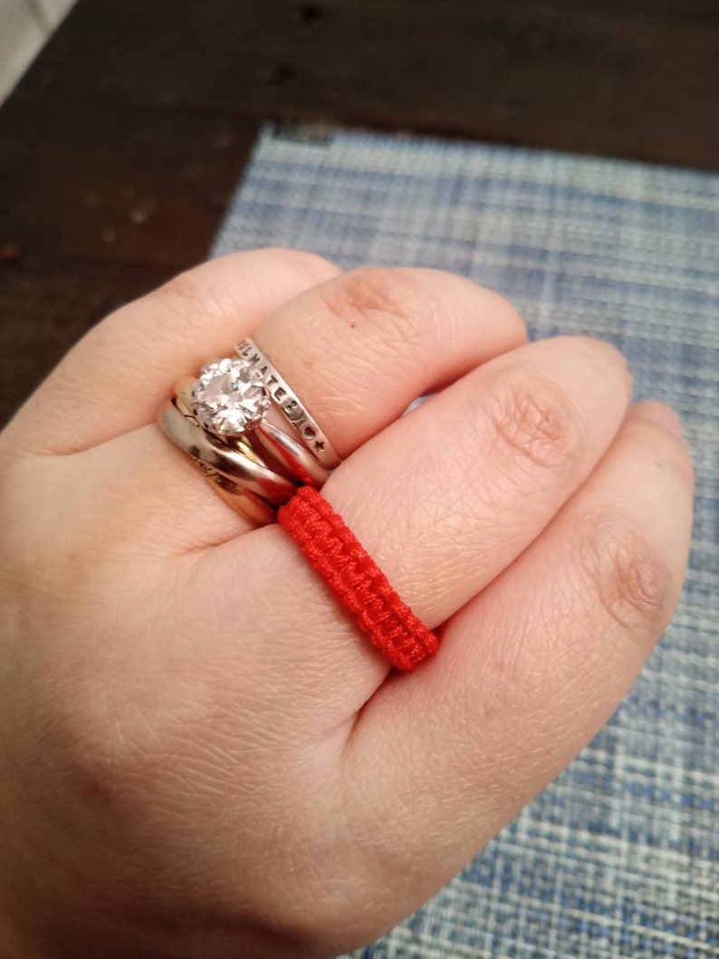 May include: A hand wearing three silver rings and a red string bracelet. The middle ring features a large, clear gemstone.