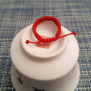 May include: A red string bracelet with a knot design. The bracelet is on a white ceramic dish with a red Chinese character.