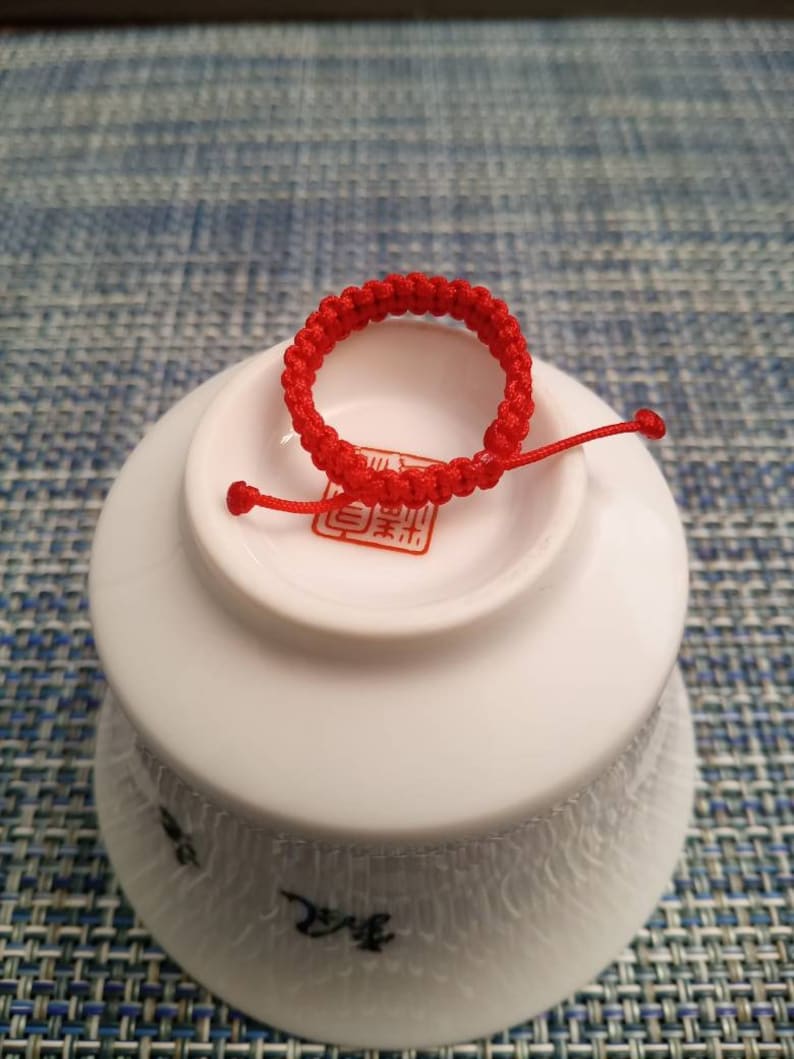 May include: A red string bracelet with a knot design. The bracelet is on a white ceramic dish with a red Chinese character.