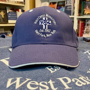 Könnte beinhalten: Eine blaue Baseballkappe mit weißem Schirm und einem weißen gestickten Logo mit der Aufschrift "Holy Cross Monastery Est. 1902 West Park, New York".