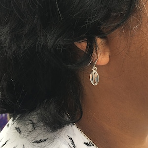 May include: Silver dangle earrings with a small, oval-shaped charm. The charm has a textured, organic design.