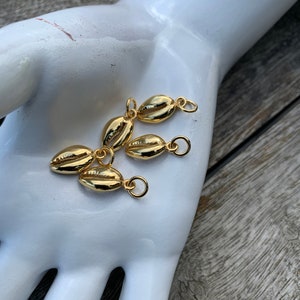 May include: Five gold-colored shell charms with jump rings. The charms are arranged in a cluster on a white surface.