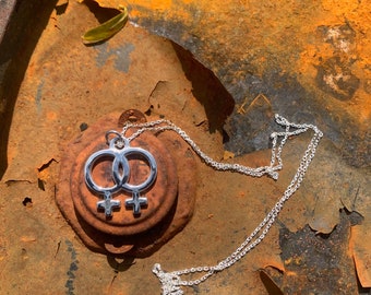 May include: A silver necklace with a double female symbol pendant. The necklace is lying on a rusty metal surface.
