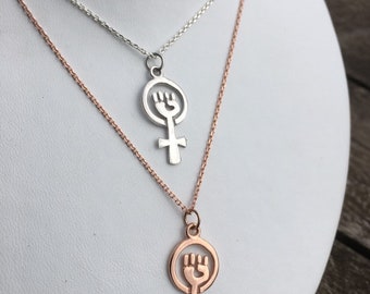 May include: Two necklaces, one with a silver finish and the other with a rose gold finish. Both necklaces feature a pendant with a raised fist inside a circle, representing the female gender symbol.