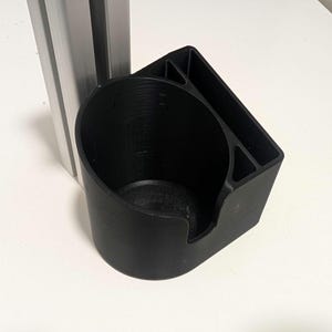 May include: A black, 3D-printed cup holder with a cylindrical cup section and a right-angled support. The holder is designed to attach to a vertical metal support. The cup holder is made of black plastic.