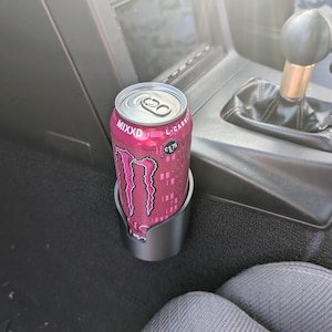 May include: A silver car cup holder holding a pink and black Monster energy drink can. The can has the word "MIXXD" printed on it. The cup holder is in a car interior.