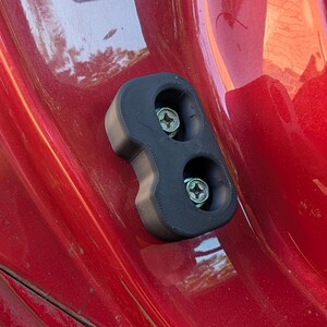 May include: A black, 3D-printed plastic component with two screw holes, attached to a red surface. The screws are silver with a star-shaped head. The part is likely a component or bracket for a vehicle or similar application.