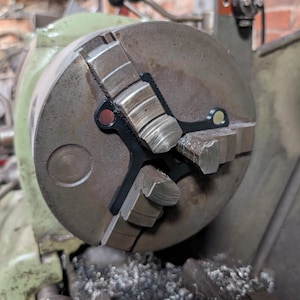 May include: A close-up shot of a metal lathe chuck with three jaws. The chuck is a dull silver colour, with black rubber inserts. The lathe is painted green and has various levers and handles. Metal shavings are visible at the base of the machine.