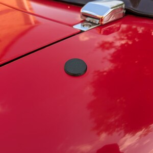 May include: A close-up shot of a vibrant red car bonnet featuring a black circular cap and a chrome latch. The bonnet reflects the sky and surroundings, giving a glossy look. The car's surface is smooth and clean.