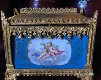 May include: A gold-colored, ornate box with a blue and white porcelain panel depicting two cherubs playing music. The box has a gothic-style design with intricate details.