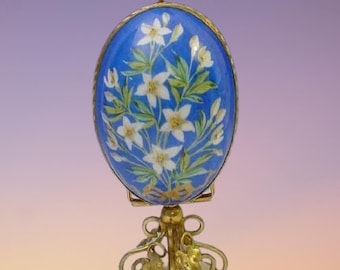 Op de afbeelding: Een blauw en gouden decoratief ei met witte bloemen erop geschilderd. Het ei staat op een gouden standaard met een witte basis.