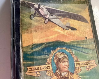 Może przedstawiać: Okładka starej książki z zielonym tłem, na której znajduje się czarno-biała ilustracja samolotu w locie. Tytuł książki to "The Boy's Story of Lindbergh The Lone Eagle". Okładka zawiera również portret Charlesa Lindberga z napisami "Clean Living", "Courage" i "Preparedness" wokół portretu.