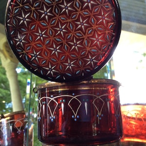 Puede incluir: Un tarro de vidrio rojo con un borde dorado y un patrón decorativo de estrellas blancas y puntos azules. El tarro tiene una tapa con un patrón similar y se asienta sobre tres patas de metal dorado.