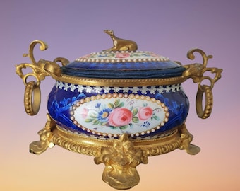 May include: A blue and white porcelain box with gold accents and a floral design. The box is decorated with a small dog figurine on the lid and sits on a gold stand with three legs.