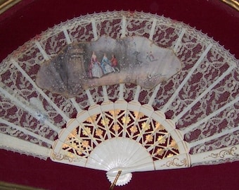May include: A white lace fan with a painted scene of people in a garden. The fan is closed and has a gold-painted design on the handle.