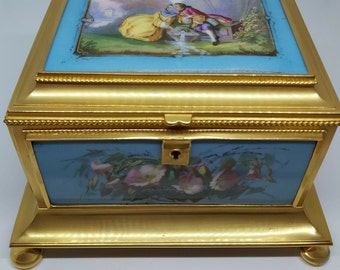 Op de afbeelding: Een met goud afgewerkte, blauw-witte porseleinen doos met een geschilderd tafereel van een stelletje bij een fontein op de deksel. De doos heeft een bloemmotief aan de voorkant en rust op vier pootjes.