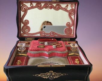 May include: An antique wooden jewelry box with a red velvet interior and a mirror in the lid. The box is decorated with gold trim and intricate floral designs. The box is open and contains various small items, including a thimble, scissors, and a small bottle.