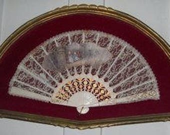 May include: A white lace fan with a gold and white painted design on the back. The fan is framed in a gold-colored frame with a red velvet backing.