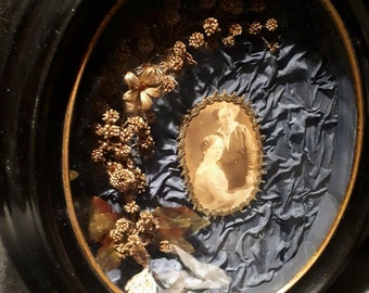 May include: A framed oval-shaped piece of art with a blue satin background. A small oval-shaped photo of a couple is in the centre of the frame. The photo is surrounded by a gold trim. The background is decorated with dried flowers and leaves.