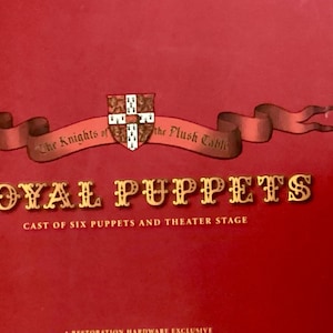May include: A red box with a gold crest and the words "The Knights of the Plush Table" and "ROYAL PUPPETS" in gold lettering. The text below reads "Cast of Six Puppets and Theater Stage".